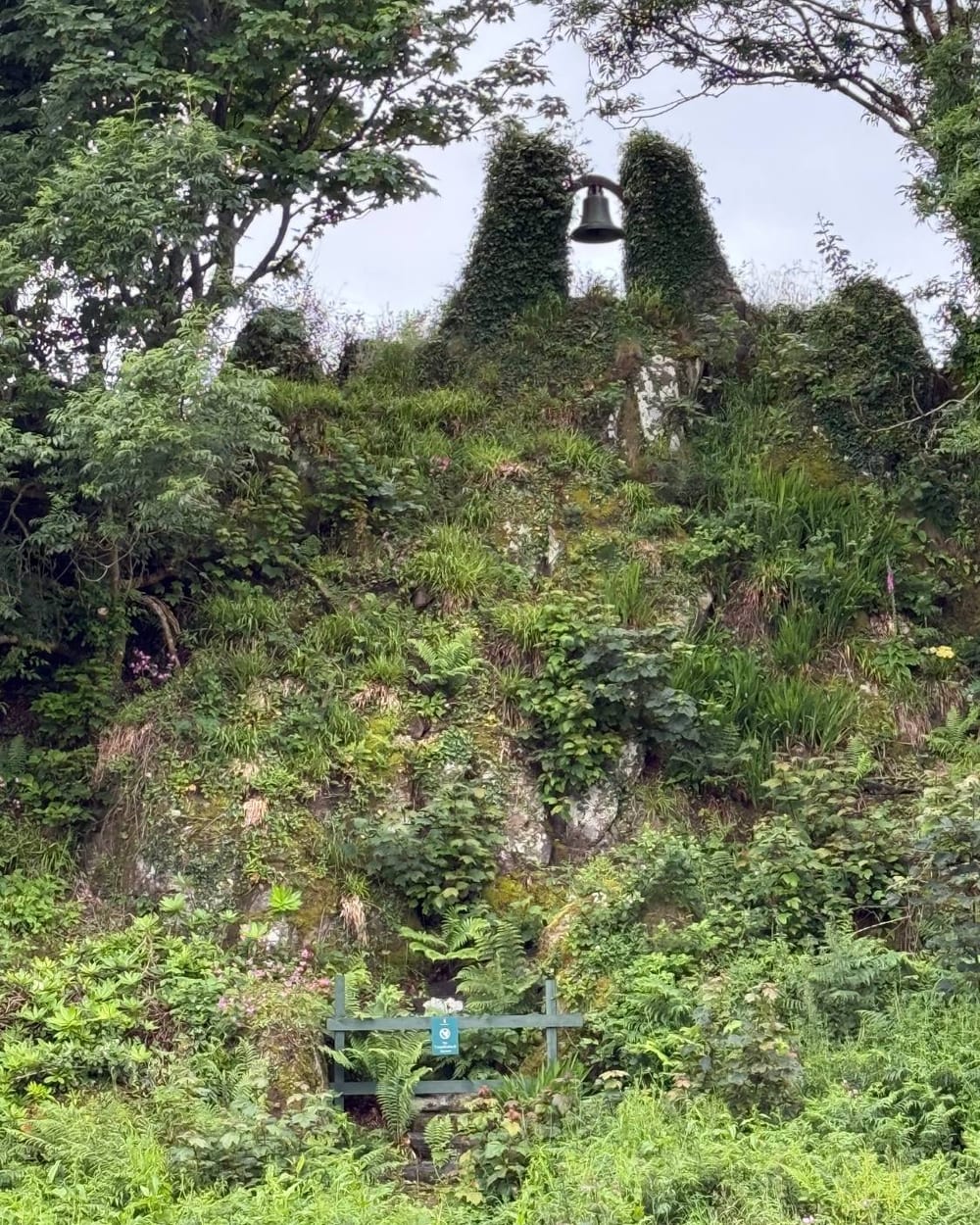 A ruined bell tower overgrown with greenery, its bell still hanging